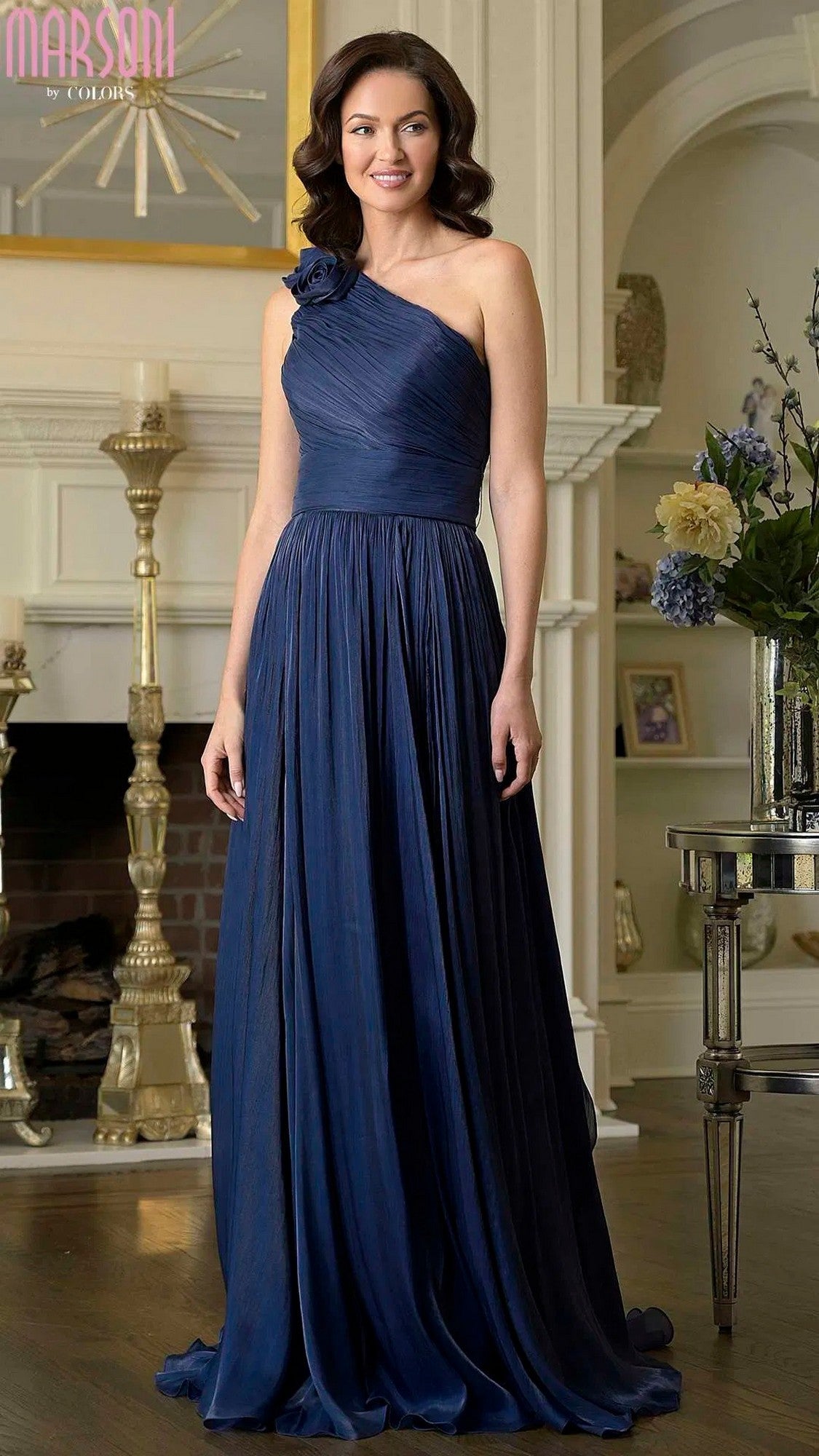 A woman in the navy Marsoni M332 long formal dress, featuring a floor-length, one-shoulder design with 3D floral embellishments, stands indoors by a fireplace and decorative shelves.